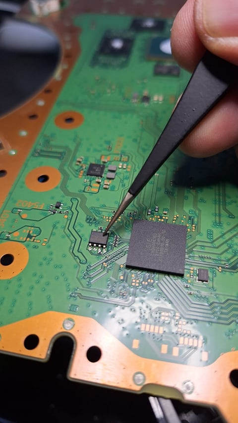 Close-up of a technician using a black tweezers to work on a green circuit board with electronic components