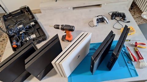 Collection of electronics and tools organized on a work surface, including gaming controllers, power drills, monitors, and various accessories on a blue mat.